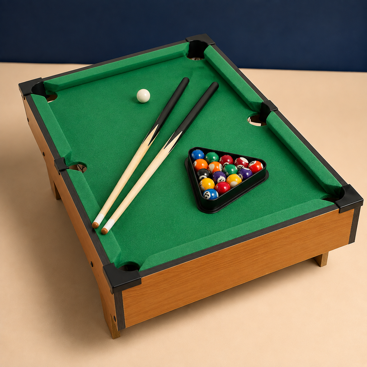 A miniature wooden billiards table with green felt, black rails, a triangle rack of colorful pool balls, a white cue ball, and two cue sticks, photographed on a beige surface against a dark blue backdrop.