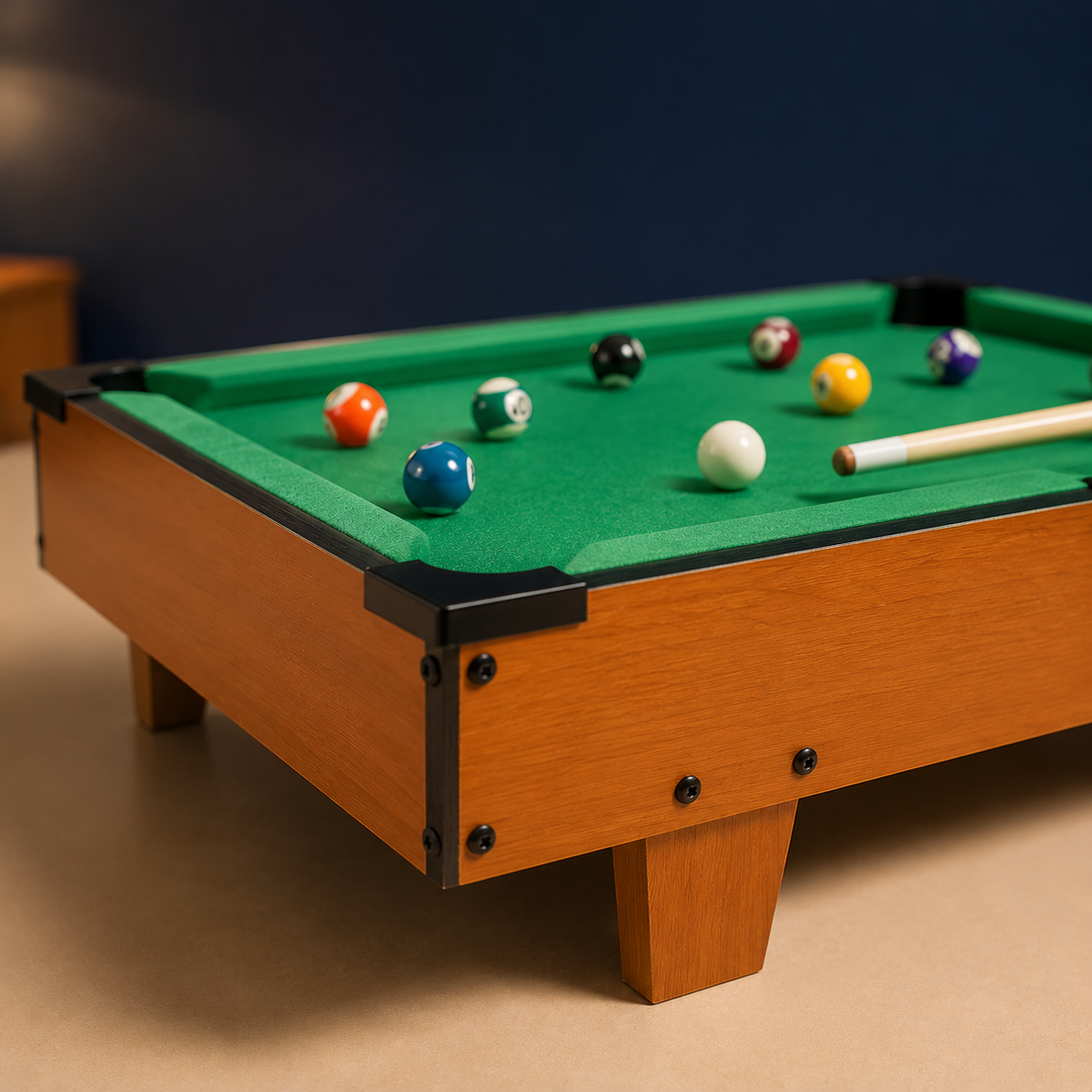 A miniature wooden pool table with a green felt surface, black corner bumpers, and several colorful billiard balls scattered on top, alongside a wooden cue stick, set against a dark blue wall and beige floor.