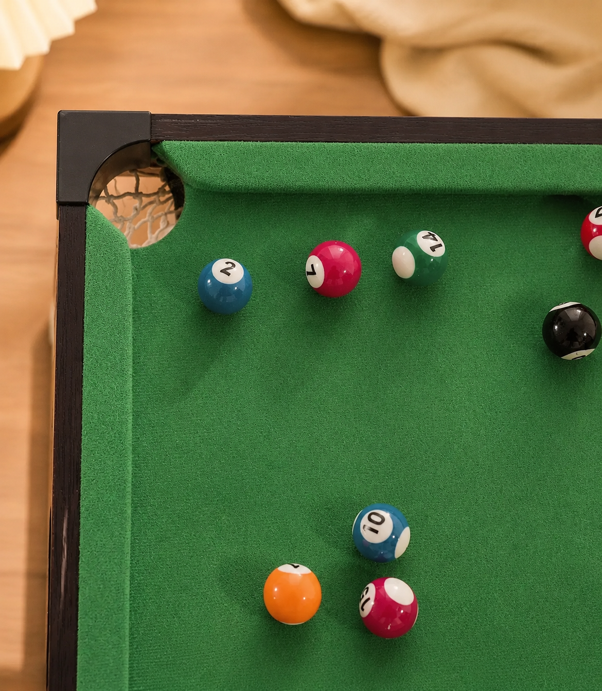 Close-up of a green felt pool table corner with several billiard balls scattered on the surface, photographed indoors on a wooden surface.